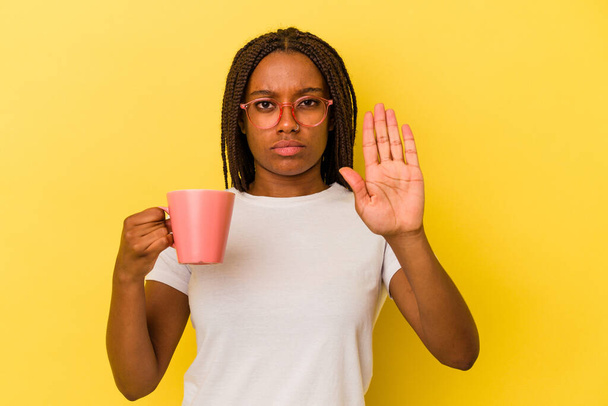 Fiatal afro-amerikai nő kezében egy bögre elszigetelt sárga háttér álló kinyújtott kézzel mutató stop jel, megakadályozza, hogy. - Fotó, kép