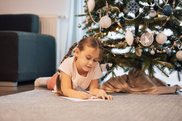 little child girl writes letter Santa Claus and dreams of a gift background New Year tree indoors. Merry Christmas and Happy Holidays. - Fotoğraf, Görsel