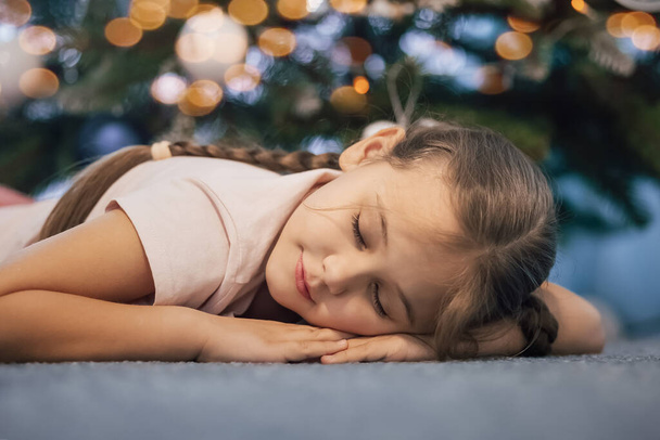 Close up shot of attractive small girl lies on floor, sleeps near New Year or Christmas tree, has pleasant dreams in evening. Children, winter holidays, rest concept - Foto, imagen