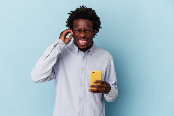 Jonge Afro-Amerikaanse man houdt mobiele telefoon geïsoleerd op blauwe achtergrond bedekt oren met handen. - Foto, afbeelding