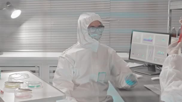 Medium view through glass wall in research center of two scientists wearing hazmat suits, gloves and medical masks, sitting at desk, working with chemicals in test tubes - Záběry, video