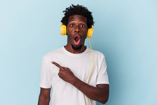 Jeune homme afro-américain écoutant de la musique isolée sur fond bleu pointant vers le côté - Photo, image