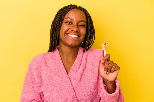 Fiatal afro-amerikai nő visel fürdőköpeny kezében borotvapenge elszigetelt sárga háttér nevetés és szórakozás. - Fotó, kép