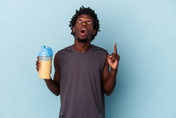 Nuori afrikkalainen amerikkalainen mies pitelee proteiinia pirtelö eristetty sinisellä taustalla osoittaa ylösalaisin suu auki. - Valokuva, kuva