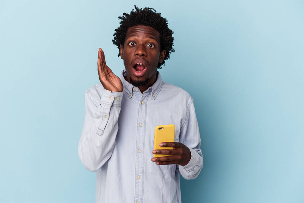 Νεαρός Αφροαμερικάνος που κρατά το κινητό τηλέφωνο απομονωμένο σε μπλε φόντο έκπληκτος και σοκαρισμένος. - Φωτογραφία, εικόνα