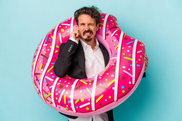 Jovem homem de negócios com donut inflável isolado no fundo azul cobrindo orelhas com as mãos. - Foto, Imagem
