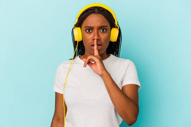 Joven mujer afroamericana escuchando música aislada sobre fondo azul guardando un secreto o pidiendo silencio. - Foto, Imagen