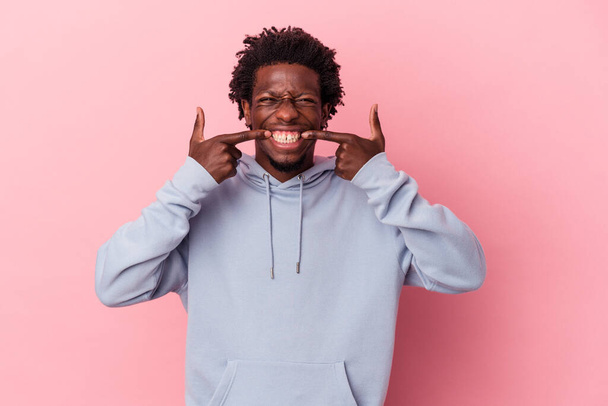 Young african american man isolated on pink background smiles, pointing fingers at mouth. - 写真・画像