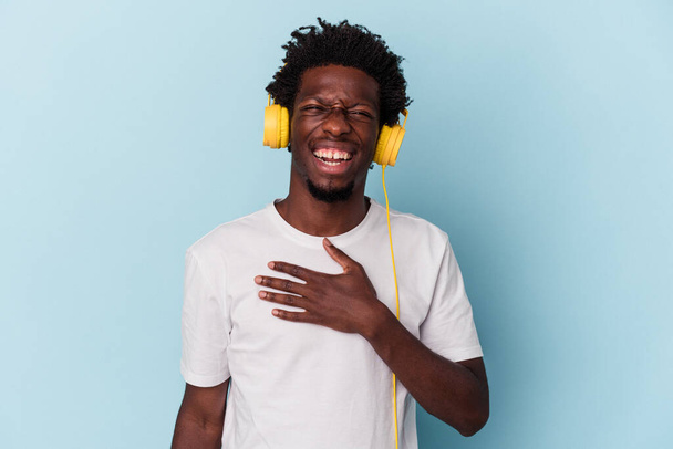 Joven afroamericano escuchando música aislada sobre fondo azul se ríe en voz alta manteniendo la mano en el pecho. - Foto, imagen