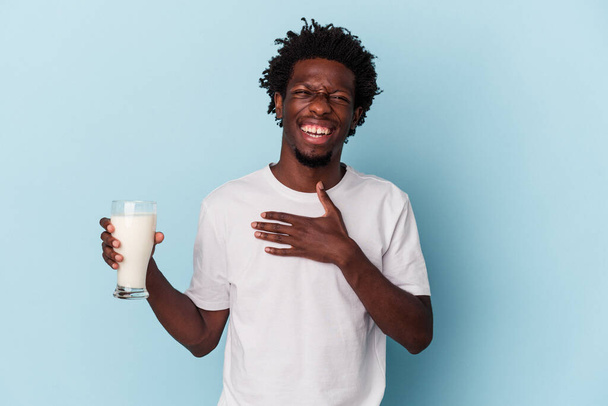 Junger afrikanisch-amerikanischer Mann hält ein Glas Milch isoliert auf blauem Hintergrund und lacht laut heraus, die Hand auf der Brust haltend. - Foto, Bild