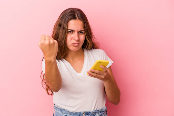 Молодая белая женщина с мобильным телефоном на розовом фоне показывает кулак к камере, агрессивное выражение лица. - Фото, изображение