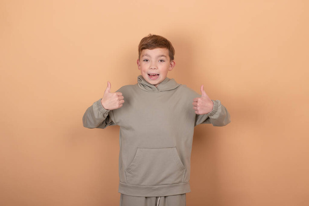 Portrait de garçon adolescent mignon heureux en vêtements verts debout et montrant geste pouce vers le haut, isolé sur fond brun - Photo, image