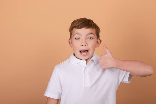 Things are good. Pretty school boy in white t-shirt looking at the camera and showing thumbs up gesture on light brown background. Portrait of handsome teenager guy 9-11 years old with like sign - Фото, зображення