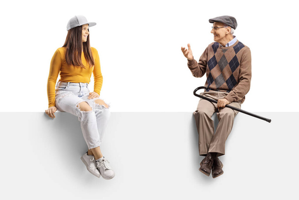 Young female and an elderly man sitting on a blank panel and having a conversation isolated on white background - Foto, immagini