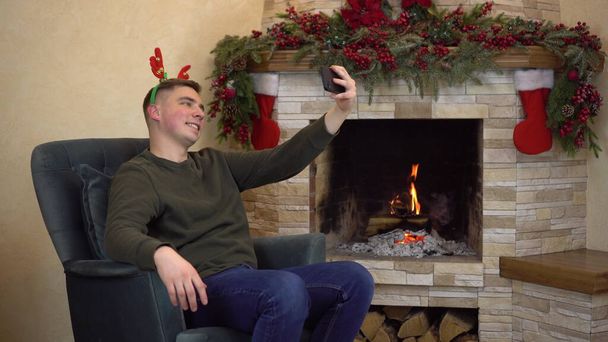 A young man sits in an armchair by the fireplace with antlers on his head and takes a selfie. Christmas mood. - Photo, Image