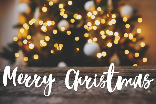 Καλά Χριστούγεννα ευχετήρια κάρτα. Καλά Χριστούγεννα κείμενο για ρουστίκ ξύλο κατά χριστουγεννιάτικο δέντρο φωτίζει χρυσό bokeh. Ατμοσφαιρική παραμονή Χριστουγέννων. Καλές γιορτές! Χειρόγραφο σήμα - Φωτογραφία, εικόνα