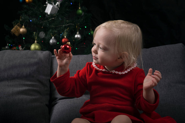 bambina si siede su un divano sullo sfondo di un albero di Natale e guarda un giocattolo dell'albero di Natale. Anno nuovo. - Foto, immagini