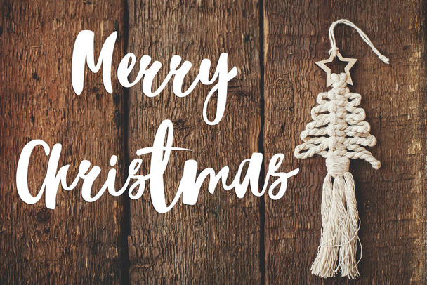 メリークリスマスグリーティングカード。木材上のクリスマスツリーのマクラメの装飾にメリークリスマスのテキストは、最小限の素朴なフラットレイアウト。ボー環境に優しいおもちゃ。ハッピー・ホリデー！手書きのサイン - 写真・画像