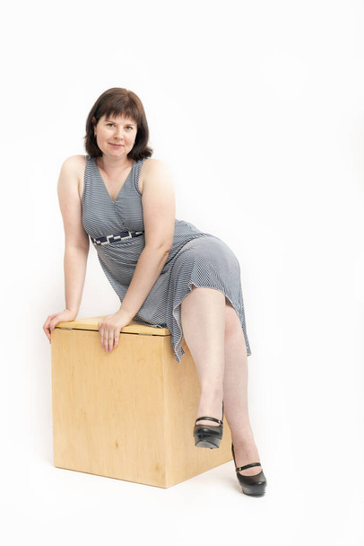 Donna di mezza età con eccesso di peso in abito seduto su una scatola di legno su sfondo bianco. Modello plus size - Foto, immagini