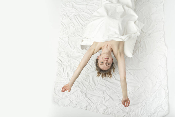 Beautiful naked woman lies in bed hands up, covered with white sheet and enjoys the weekend. Relaxing at home. White background. Top view. - Valokuva, kuva