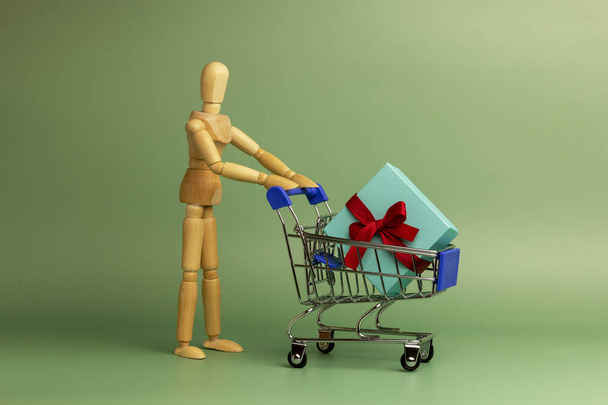 Іграшкова модель дерев'яного чоловіка з візком і подарунковою коробкою. Макет для реклами подарунків, одягу, покупок. Копіювати простір
. - Фото, зображення