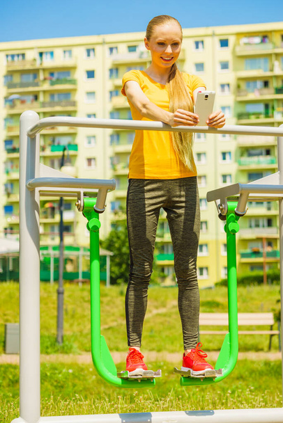 Young woman working out in outdoor gym. Girl taking selfie photo with smartphone while doing exercises on street machine. - Φωτογραφία, εικόνα