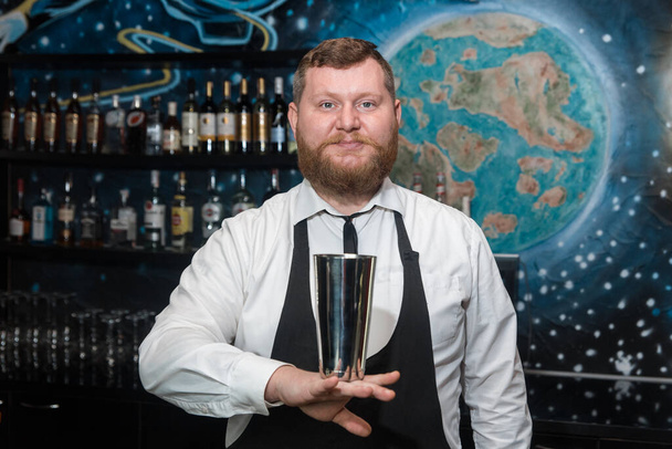 A bearded adult man of European appearance, a professional bartender, holds in his hands a tool for preparing and mixing alcoholic cocktails in a nightclub. - Photo, Image