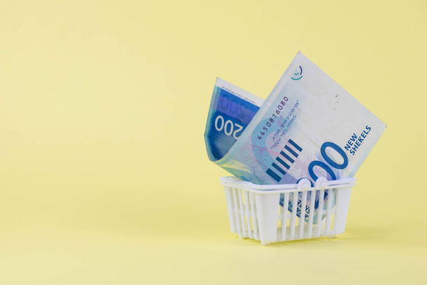 Izrael peníze šekel bankovky v nákupním košíku na žlutém pozadí. Kurzové kurzy a nákup koncepce prodejní měny. - Fotografie, Obrázek