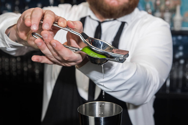 Руки профессионального бармена выжимают сок лайма железным инструментом с соковыжималкой в металлический шейкер крупным планом. - Фото, изображение