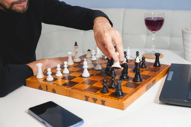 Задумчивый лысый мужчина с бородой играет в шахматы на борту рядом с ноутбуком и бокалом вина. Концепция стратегии и конкуренции. Вид сбоку - Фото, изображение