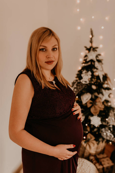 Schwangere im langen schwarzen Kleid neben dem weihnachtlich geschmückten Baum. Heiligabend. Silvester im Innenraum. Valentinstag-Feier - Foto, Bild