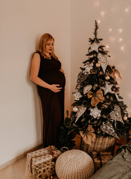 pregnant woman in long black dress near the Christmas decorated tree. Christmas morning. New Year's interior. Valentine's Day celebration - Фото, зображення