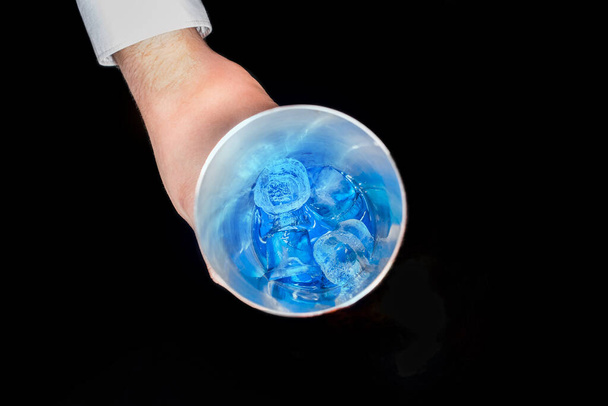 The hand of professional bartender takes a tool for mixing and making alcoholic cocktails shaker with blue cold syrup with ice in on a black background, close-up. - Photo, Image
