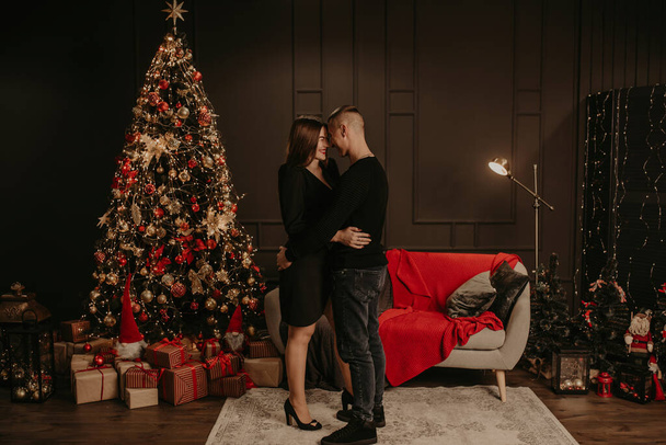 クリスマスツリーの近くでキスしてるカップルの男と女を愛してる。新年のために飾られた家。クリスマスの朝。アパートの中だ。バレンタインデーのお祝い - 写真・画像