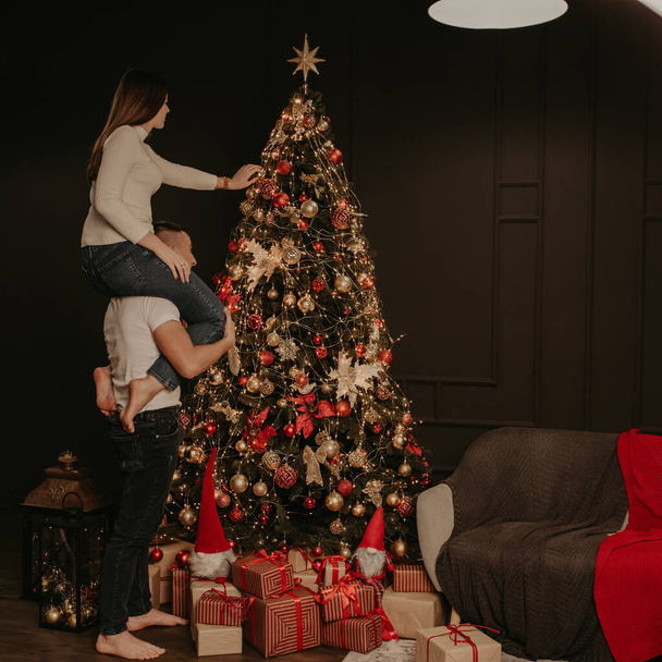 young woman sits on back shoulders of man and decorates a Christmas tree, hangs ornaments. decorated house for New Year. Christmas morning. apartment interior. Valentine Day celebration - Photo, Image