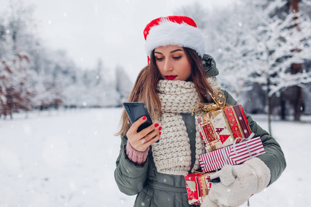 Portret van een jonge vrouw in Santa hoed met behulp van telefoon in besneeuwde winter park met een hoop kerstcadeaus buiten. Feestelijke geschenkdozen. Vrienden sms 'en via internet - Foto, afbeelding
