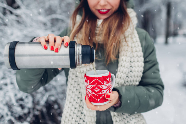 Donna che versa tè caldo dal pallone vuoto nel parco invernale innevato. Tazza con bevanda vestita con maglione o sciarpa natalizia in maglia rossa per tenerla al caldo - Foto, immagini