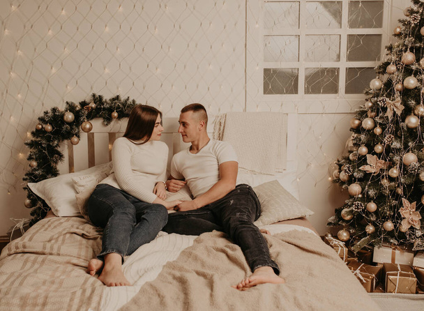 ζευγάρι άντρας και γυναίκα βρίσκονται στο κρεβάτι υπνοδωμάτιο κοντά στο χριστουγεννιάτικο δέντρο.διακοσμημένο σπίτι για τη Νέα Γενιά.Χριστούγεννα πρωί. εσωτερικό διαμέρισμα. Γιορτή του Αγίου Βαλεντίνου - Φωτογραφία, εικόνα