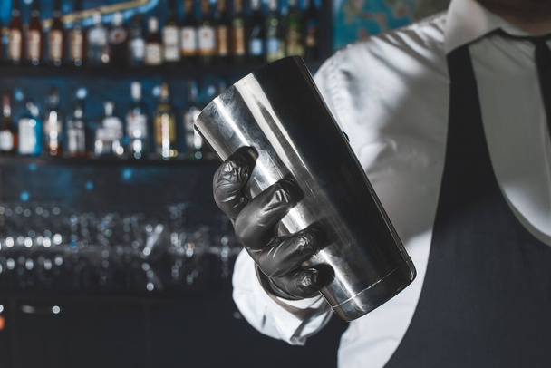 Ammattimaisen baarimikon käsi mustassa lateksikäsineessä, jossa on baari-instrumentti alkoholijuomien sekoittamiseksi ja valmistamiseksi ravistimen kanssa. - Valokuva, kuva