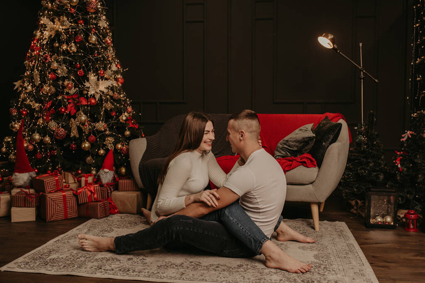 恋に二人の男と女が床に座って抱き合ってクリスマスツリーにキスする.  - 写真・画像