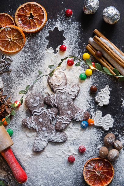 Két karácsonyi mézeskalács süti sötét háttérrel, fehér liszt, száraz narancs, fűszerek és sodrófa, lapos fektetés - Fotó, kép
