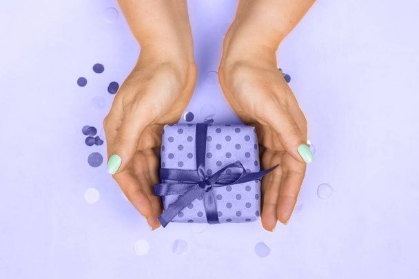 Manos de mujer con manicura pastel azul sosteniendo un regalo con un lazo sobre un fondo morado muy peri 2022. El concepto de la fiesta, regalos, sorpresas, cumpleaños, año nuevo y Navidad - Foto, imagen