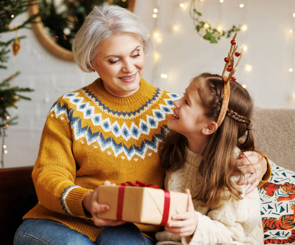 Маленька дівчинка-онучка дарує різдвяну подарункову коробку усміхненій бабусі під час зимових канікул
 - Фото, зображення