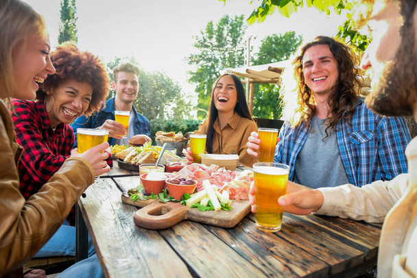Group of multiethnic happy friends living healthy lifestyle and smiling and jocking while drinking beer at outdoor pub restaurant - Young people enjoying drinks during happy hour at bar - Photo, Image