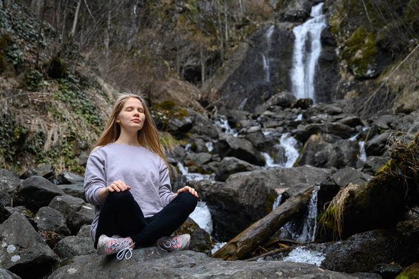 Dziewczyna siedzi na dużym kamieniu i medytuje w pobliżu wodospadu nad górską rzeką. - Zdjęcie, obraz