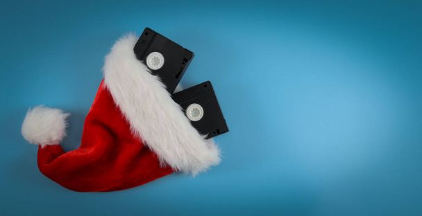 Due videocassette retrò con cappello di Babbo Natale a sinistra su sfondo blu con spazio di copia a destra, primo piano vista dall'alto. Concetto di cinema, Natale. - Foto, immagini