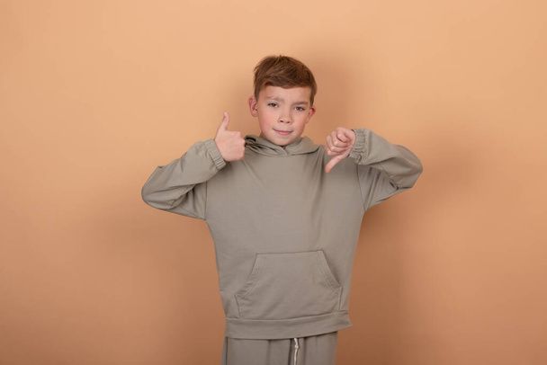 teenage handsome boy against brown studio background showing thumbs up and thumbs down, difficult choice concept - Foto, Imagen