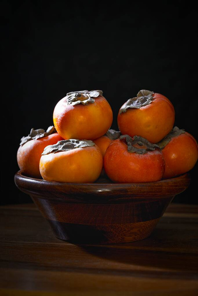 A close up still life photo of a bunch of persimmons in a wooden bowl. - Photo, Image