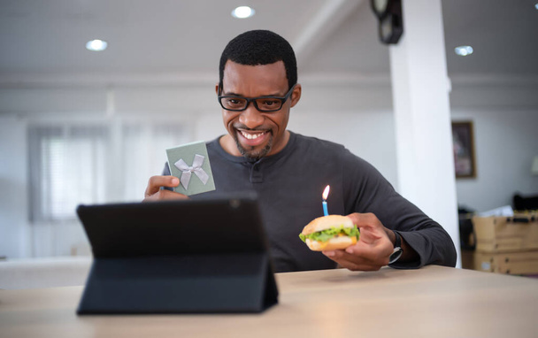 Happy Black man sitting at the table celebrating birthday through video call virtual party with friends at home. Lits and blows out candle on burger and holding gift box. Concept of distance relations - Photo, Image