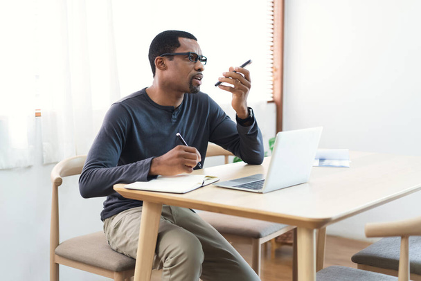 Afroamerikanisch bärtiger Freiberufler mit Laptop und Smartphone in Mundnähe, der Sprachnachrichten aufzeichnet, während er am Schreibtisch sitzt. Telefonieren über externe Lautsprecher - Foto, Bild